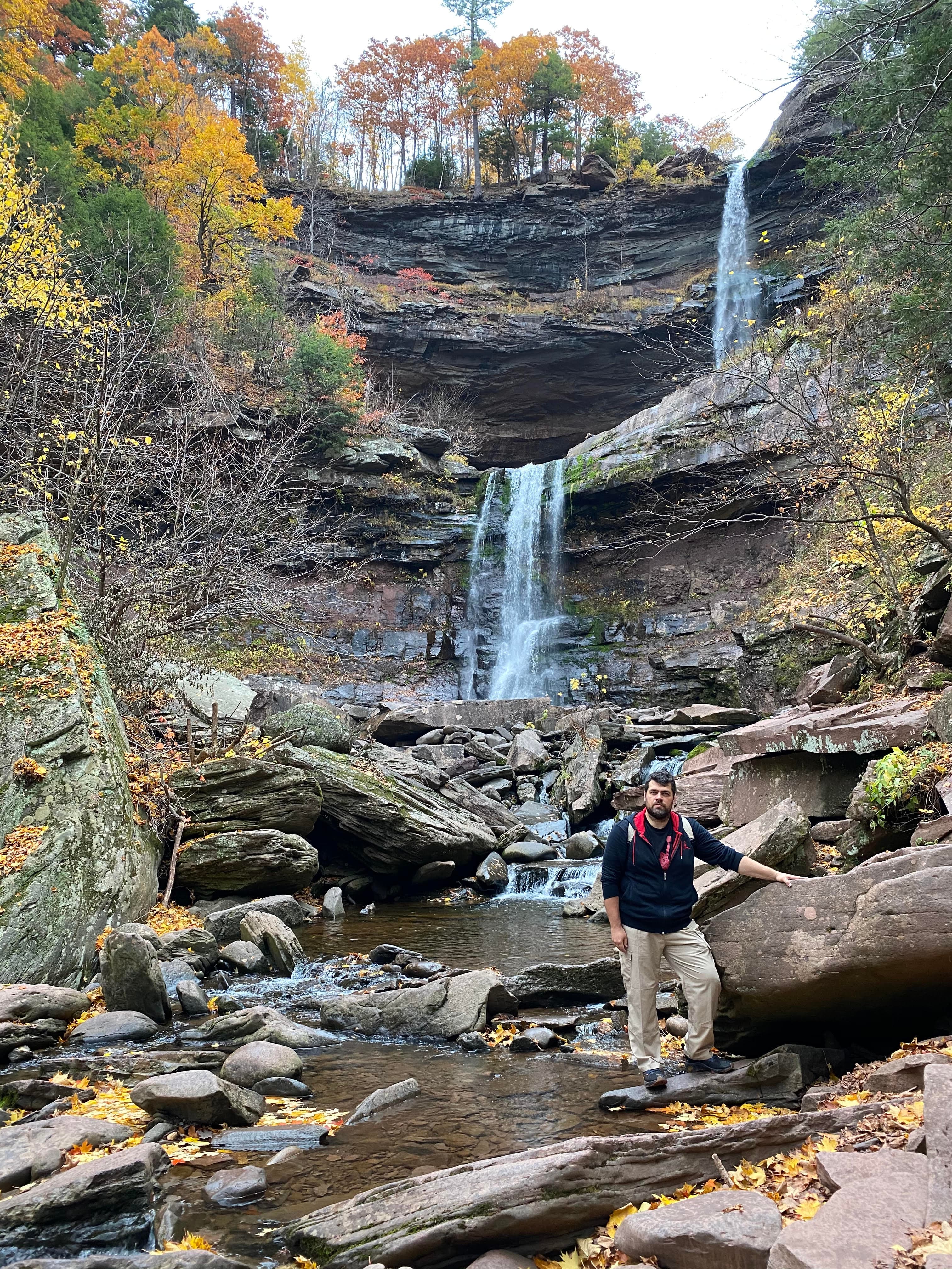Valentin standing in the lower-right beside a waterfall and layered rock, surrounded by woodland.